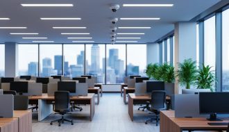 Open‑plan office with LED panel lighting, ceiling motion sensors, and floor‑to‑ceiling windows providing ample daylight, photographed straight‑on at eye level in a clean, modern setting.